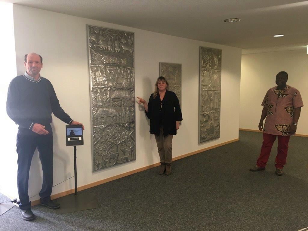 Vor des Büros der Fachstelle Inklusion und des Integrationslotsen der Stadt Bayreuth werden 2021 drei Arbeiten von Asiru Olatunde präsentiert. Margit Lebershausen, Ibukun Koussemou und Philipp Schramm stehen neben den Werken.