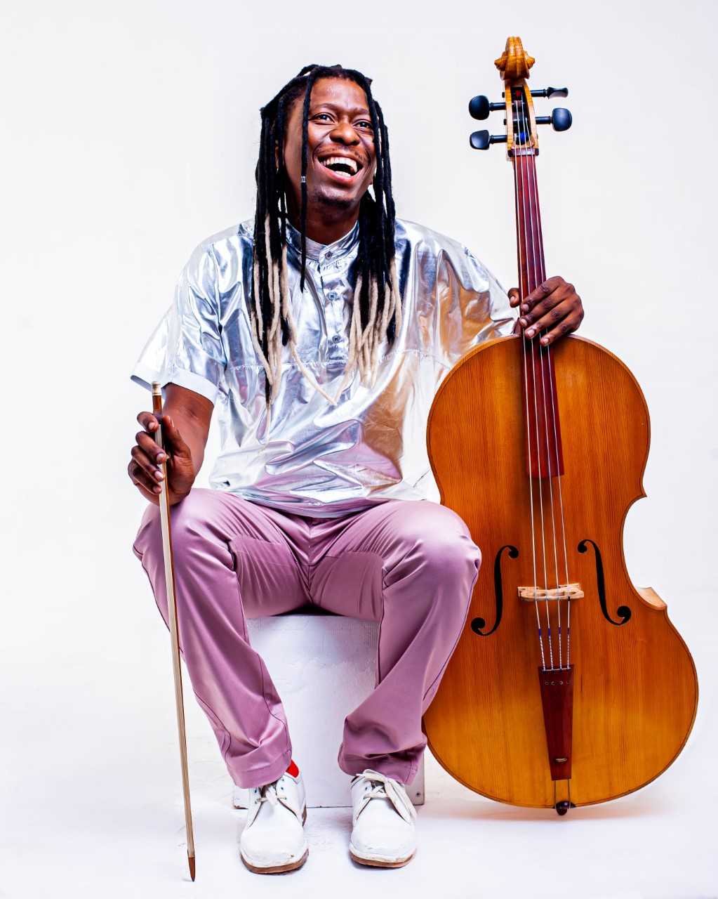 Farbfotographie: Der Künstler sitzt auf einem Stuhl, dem Betrachter gegenüber, weiße Schnürschuhe, altrosafarbene Hose, ein silbrig glänzendes Kurzarmhemd. In der rechten Hand hält er den Bogen, mit der linken Hand umgreift er den Hals eines Cellos.