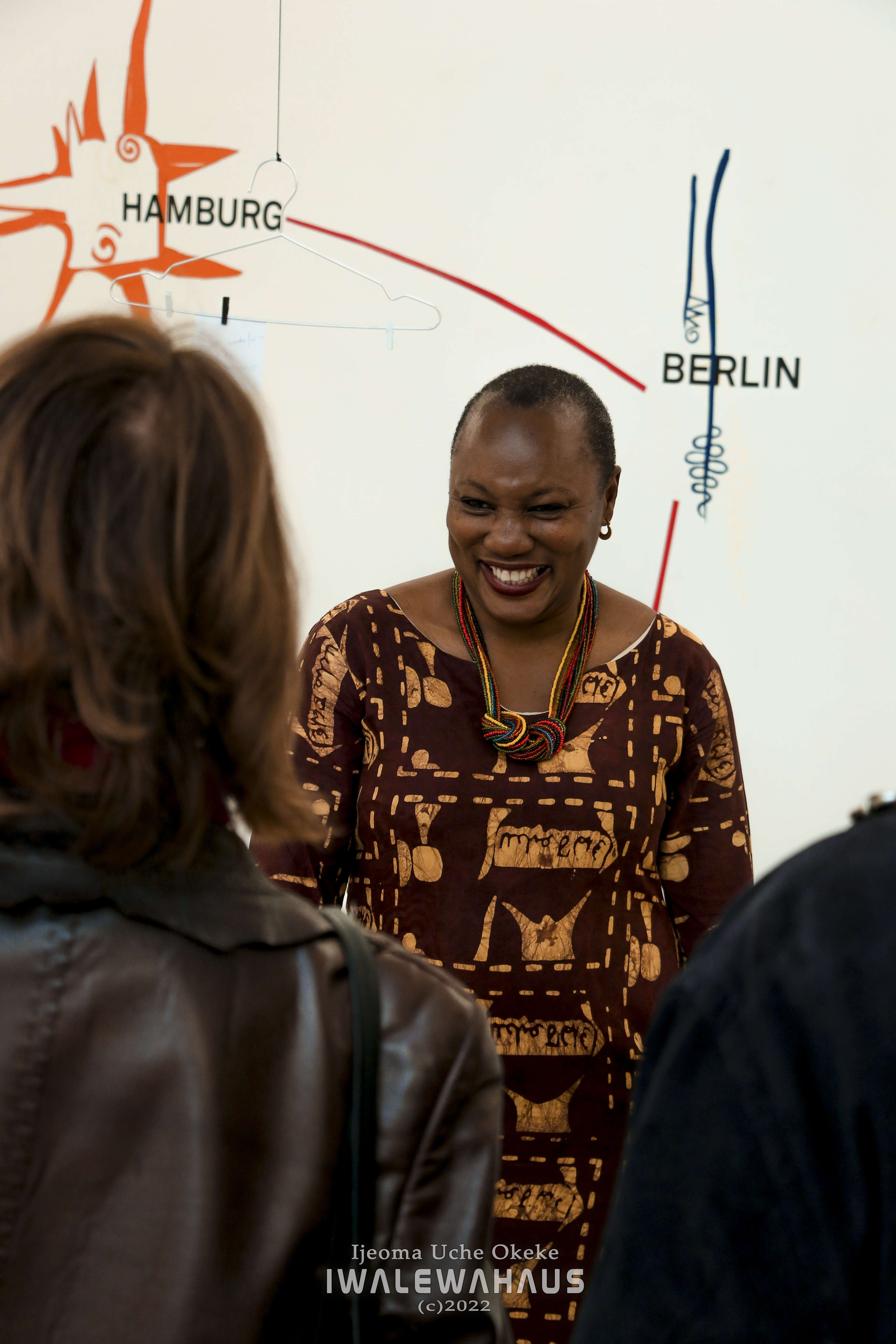 Ijeoma Uche Okeke führt durch die Ausstellung "We will now go to Kpaaza" im Iwalewahaus. Hochformatiges Farbfoto. Ijeoma Uche-Okeke trägt ein rotbraunes langes Gewand mit gelben Mustern bedruckt. Um den Hals trägt sie eine geknotete Kette.